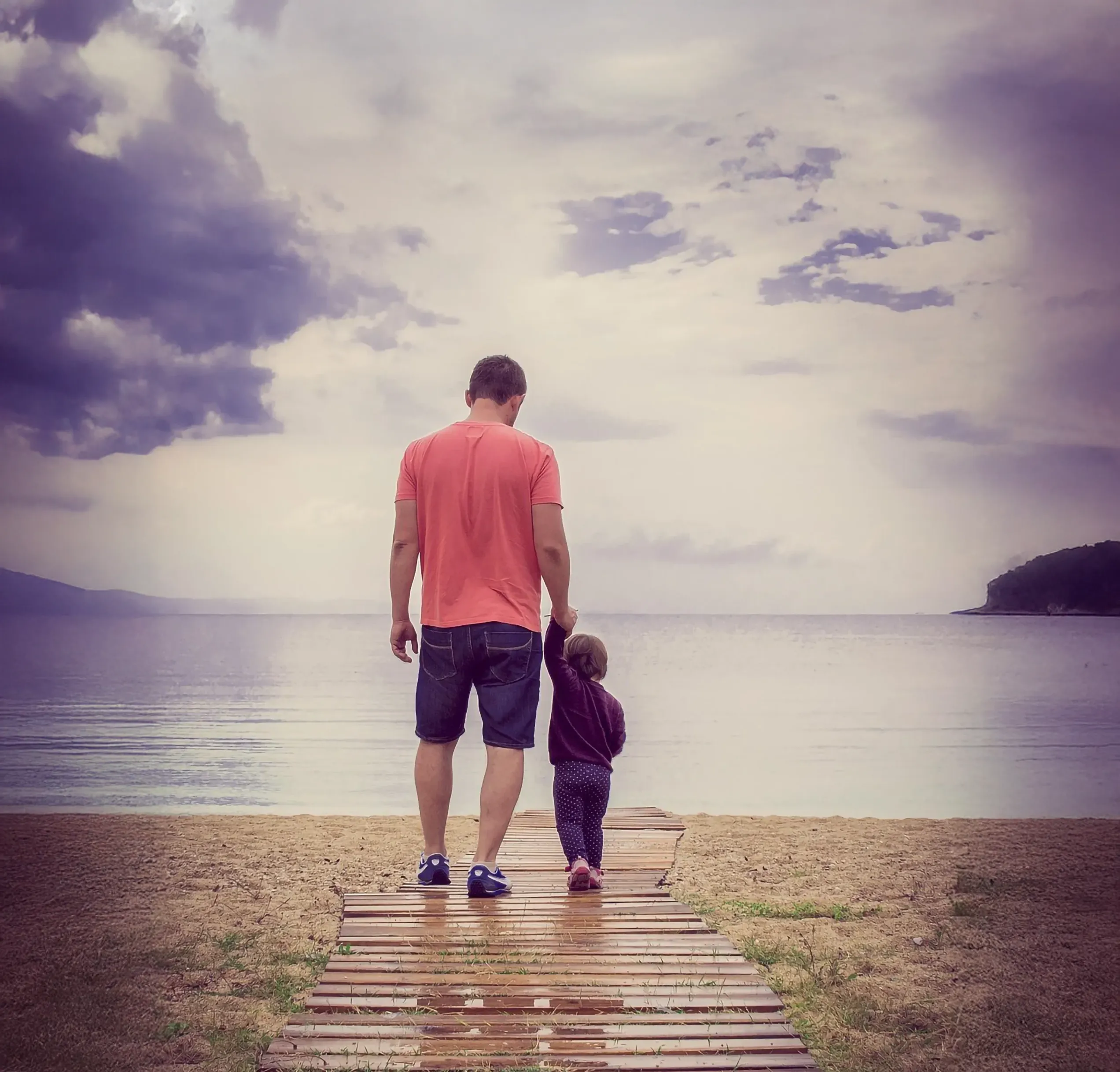 Ein Vater mit seinem Kind auf einem Holzsteg. Dahinter sieht man unscharf das Meer. Vertrauenswelt, durch Ermutigung deiner Kinder.