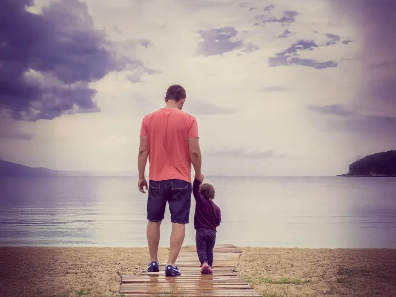 Ein Vater mit seinem Kind auf einem Holzsteg. Dahinter sieht man unscharf das Meer. Vertrauenswelt, durch Ermutigung deiner Kinder.