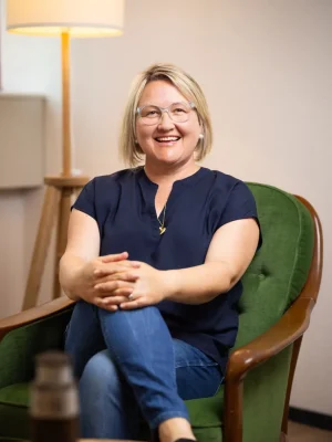 Ein Portraitbild von Thea Josi Stauber von Vertrauenswelt. Sie sitz auf einem grünen Sessel und schaut direkt in die Kamera. Der Beratungsraum, in welchem Sie sitz, ist hell und einladend.