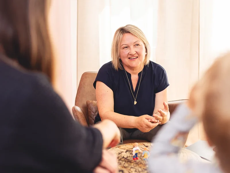 Ein Portraitbild von Thea Josi Stauber von Vertrauenswelt zwischen zwei Kunden hindurch fotografiert. Sie sitz auf einem Ledersessel und schaut die Kunden an. Der Beratungsraum, in welchem Sie sitz, ist hell und einladend.
