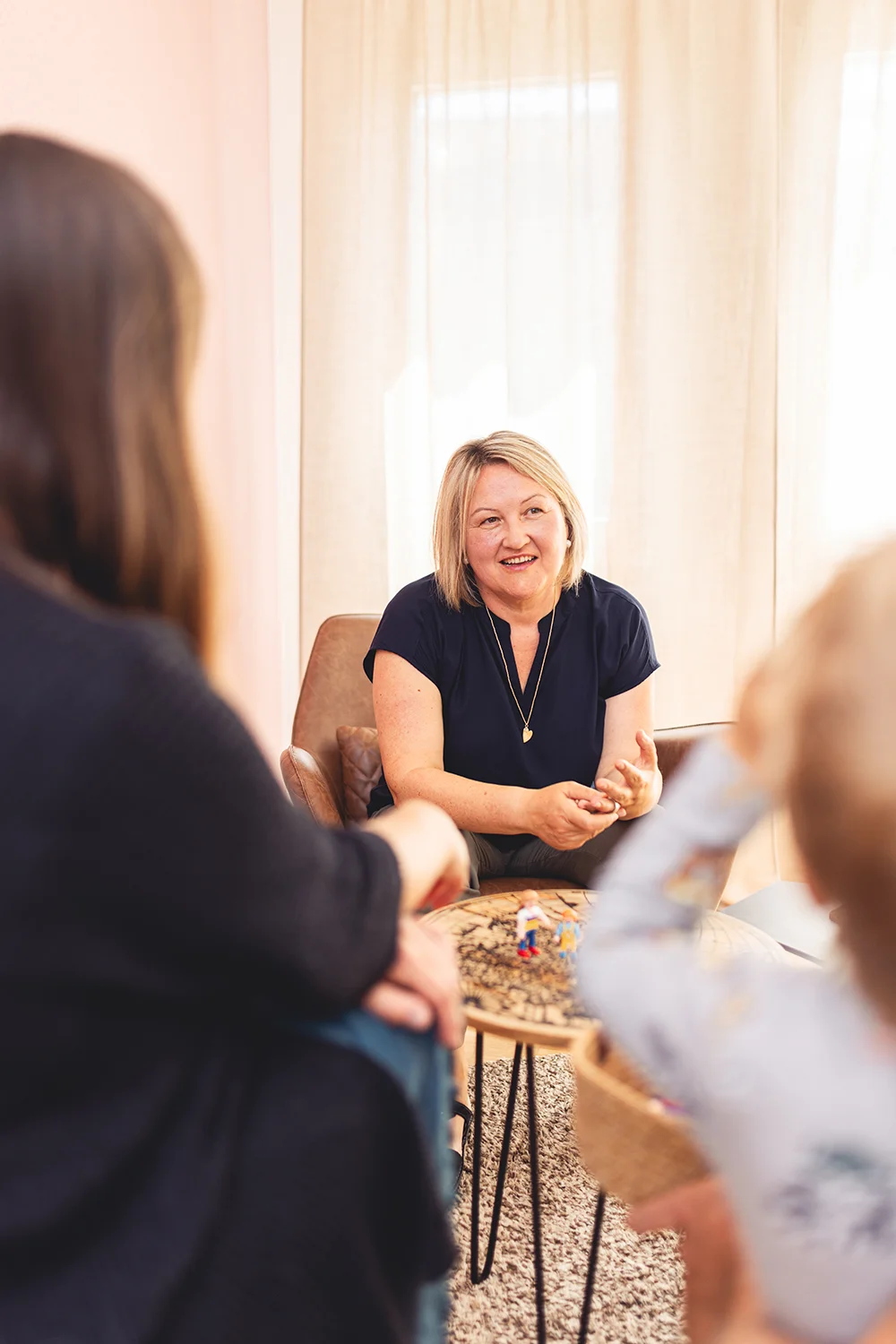 Ein Portraitbild von Thea Josi Stauber von Vertrauenswelt zwischen zwei Kunden hindurch fotografiert. Sie sitz auf einem Ledersessel und schaut die Kunden an. Der Beratungsraum, in welchem Sie sitz, ist hell und einladend.